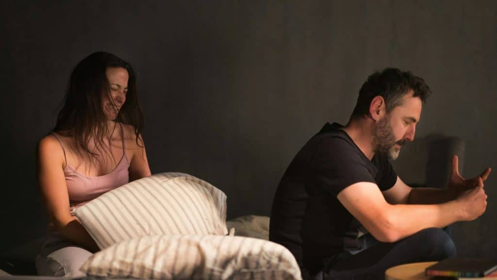 A woman holding a pillow cries, while a man next to her looks downcast.