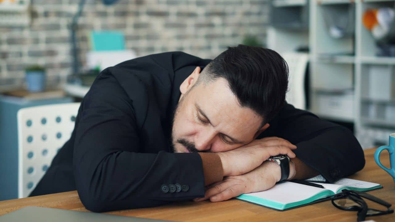 A tired man with a beard and mustache is sleeping with his head on his desk.