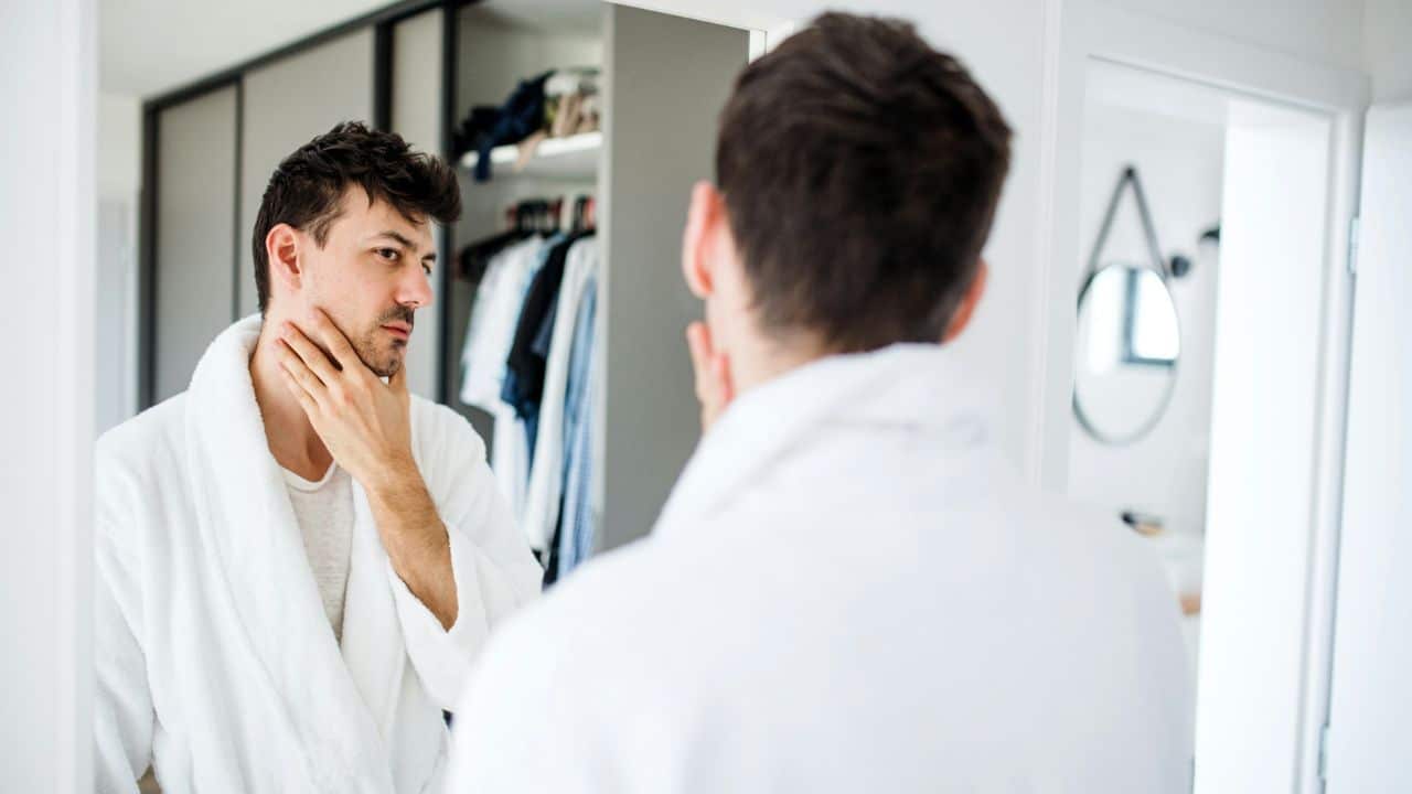 A man in a white robe looks in a mirror, touching his chin and face.