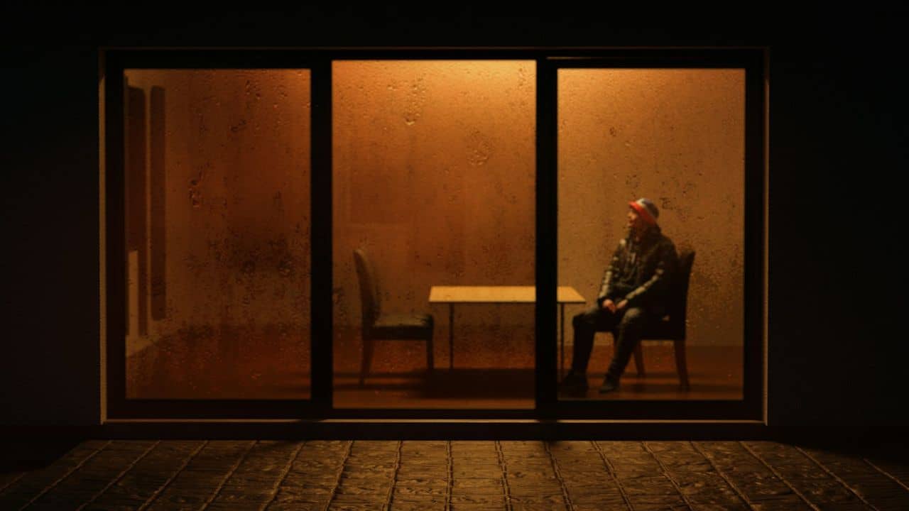 A lonely man in a jacket and baseball cap sits at a table in a dark room.