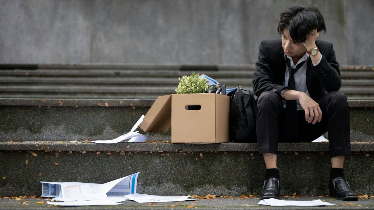 A depressed man sits on concrete stairs with a box of his belongings.