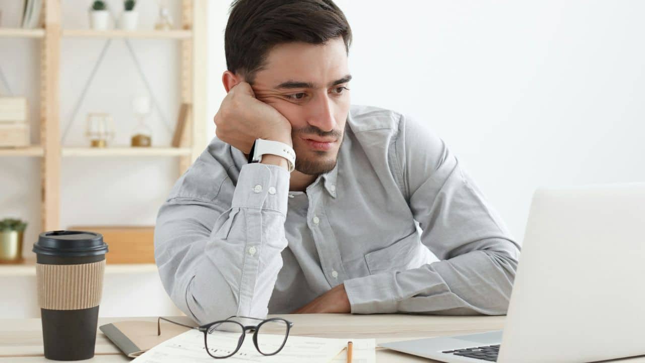 A bored man sits at a desk, resting his head on his hand and looking at a laptop.