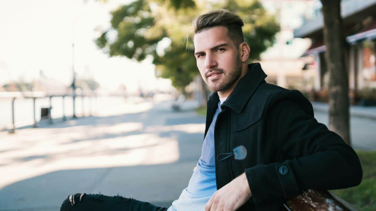 A young man with light hair and a beard sits on a bench outside.