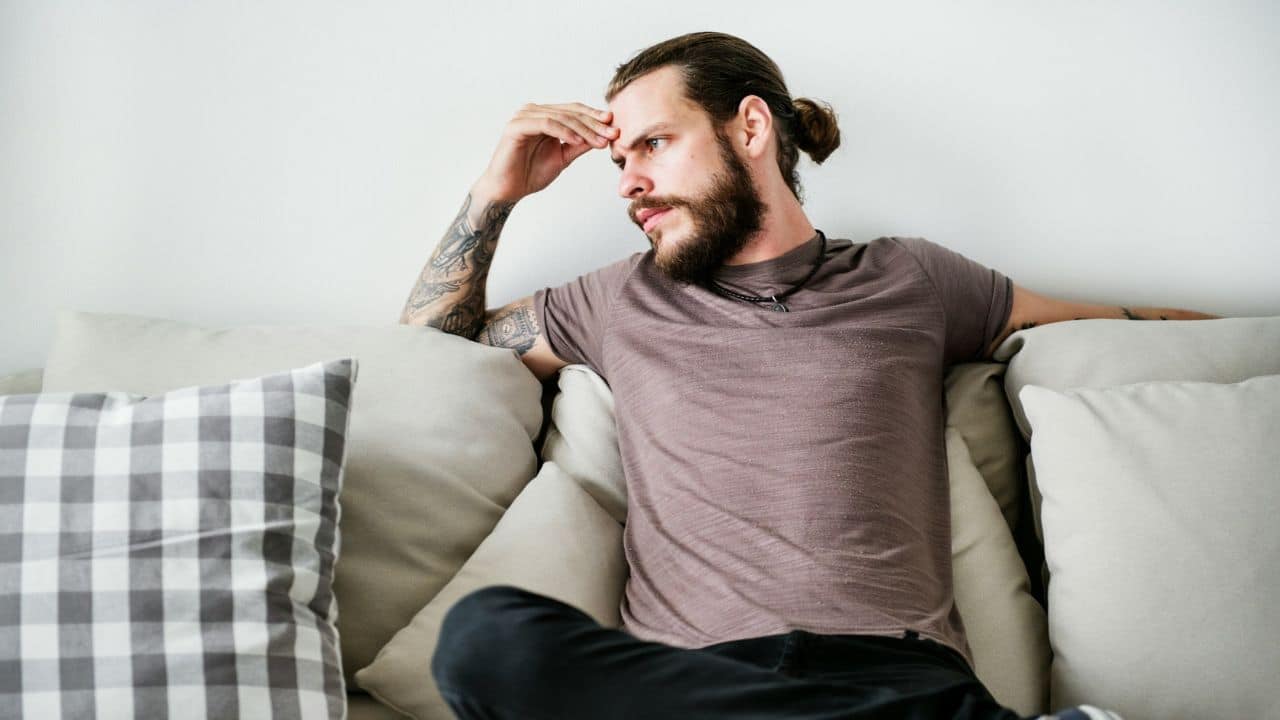 A man with a beard, man bun, and tattoos is sitting on a couch with a worried expression.