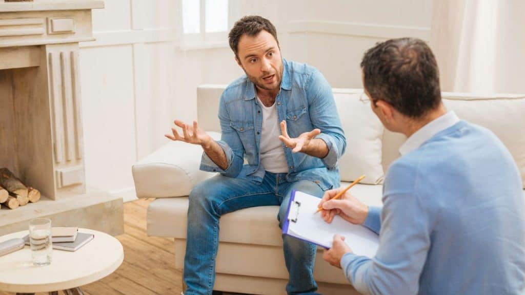 A man in a denim shirt sits on a sofa talking to a therapist who is writing on a clipboard.