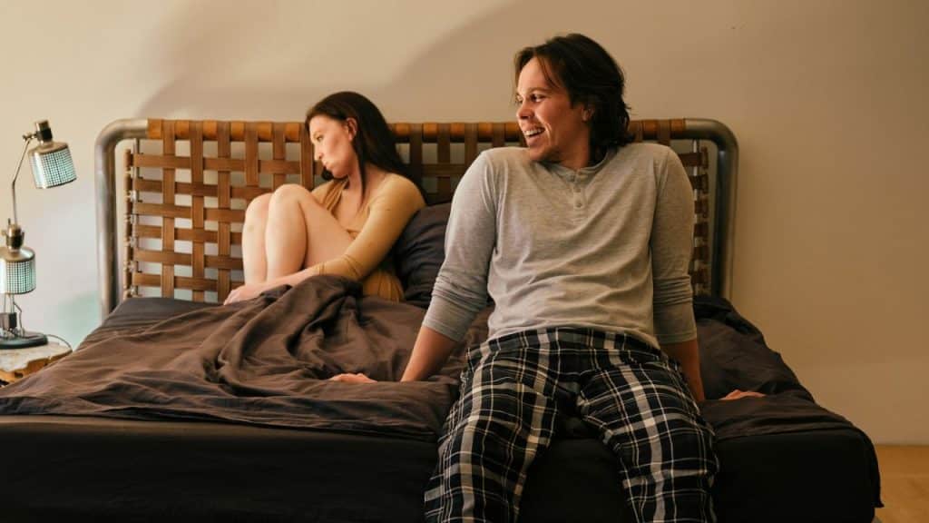 A man smiles and looks to the right while a woman looks away from him on the bed.