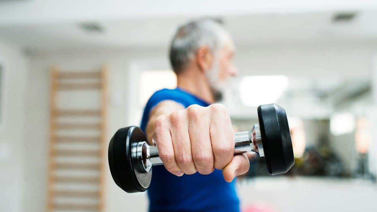 A person's hand holds a dumbbell, with a blurry background of a person exercising.