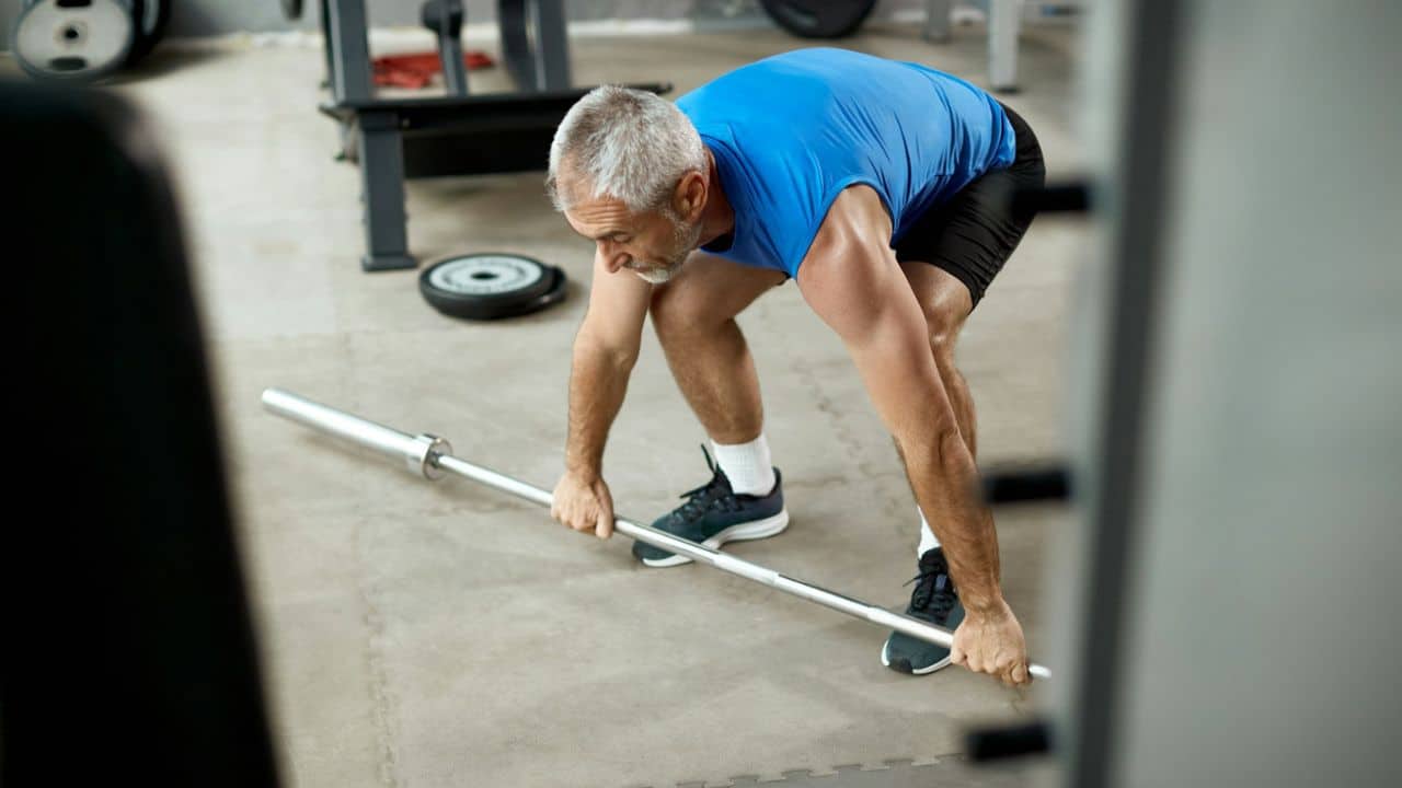 A senior man in a blue shirt and black shorts gets ready to lift a barbell.