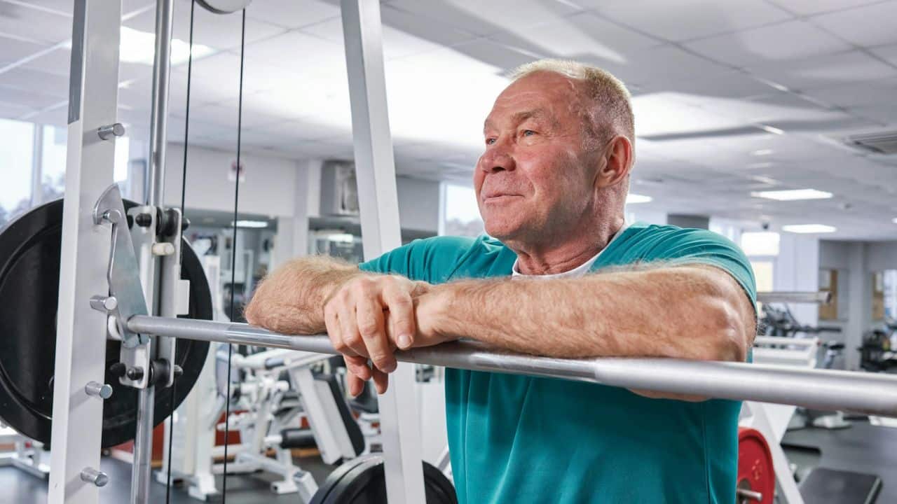 A contented-looking older man leans on a weight machine in a gym.
