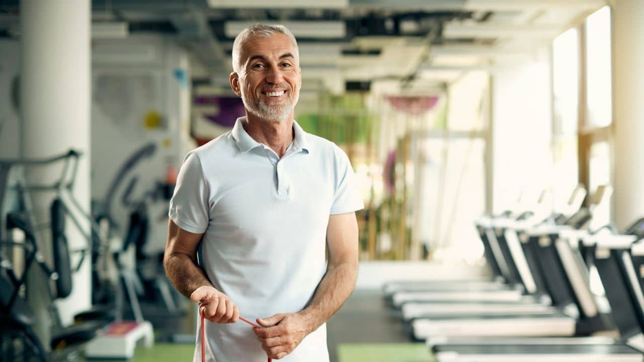 A handsome, happy, older man with a beard stands in a gym, holding a resistance band.