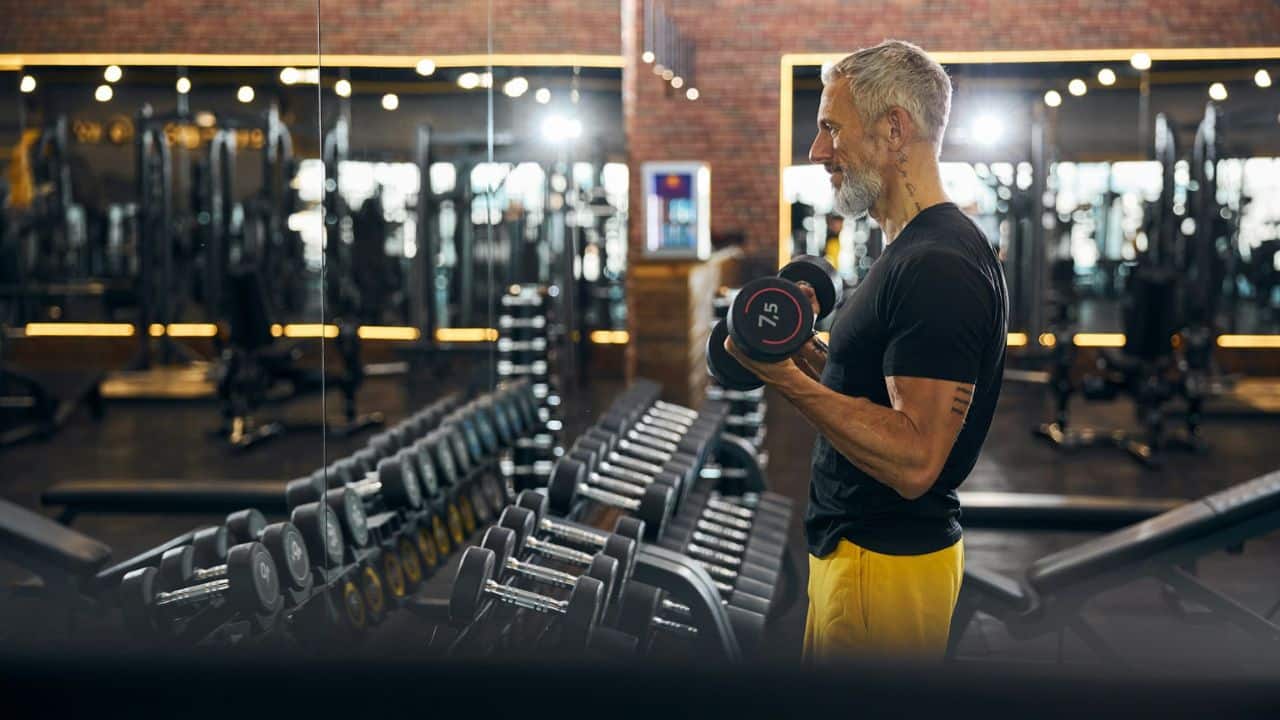 A mature, fit man with a grey beard lifts dumbbells in a well-equipped gym.