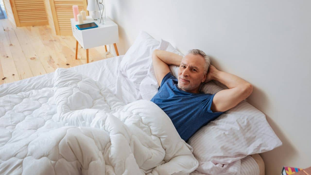 A content older man with grey hair is lying in bed with his hands behind his head.