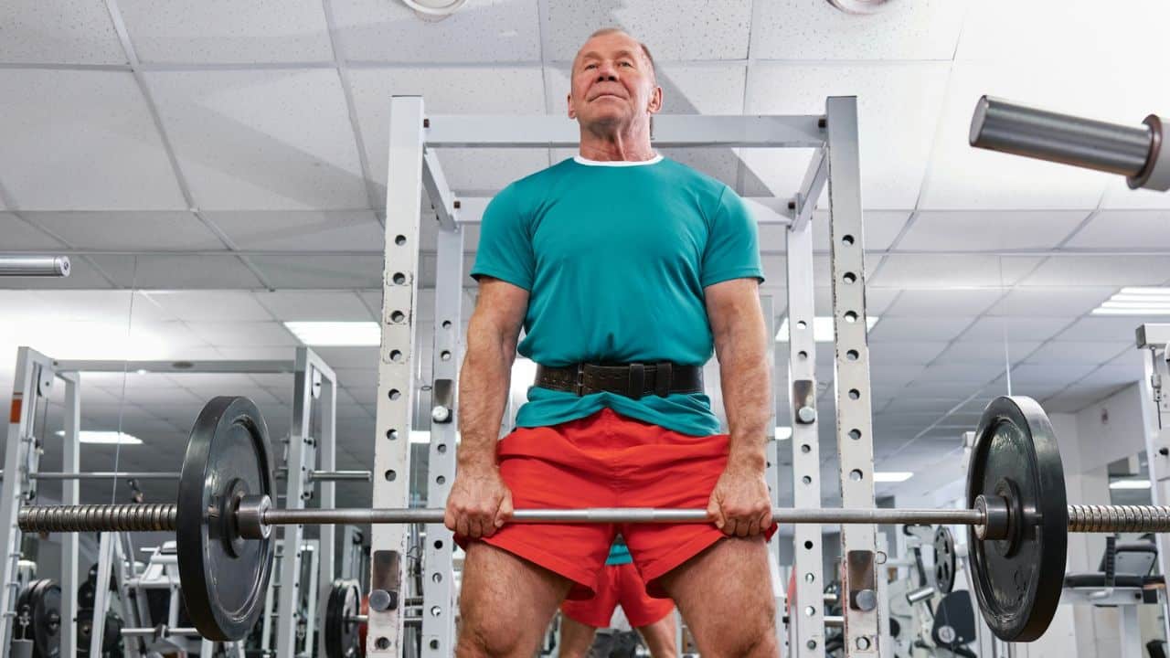 A senior man in a turquoise shirt and red shorts lifts a barbell, with a determined look on his face.