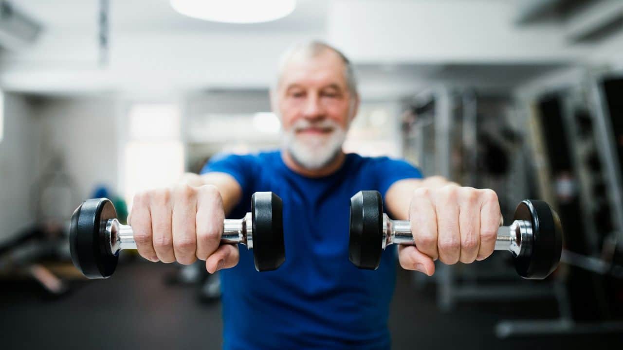 A blurred older man smiles in a gym, holding two dumbbells out in front of him.