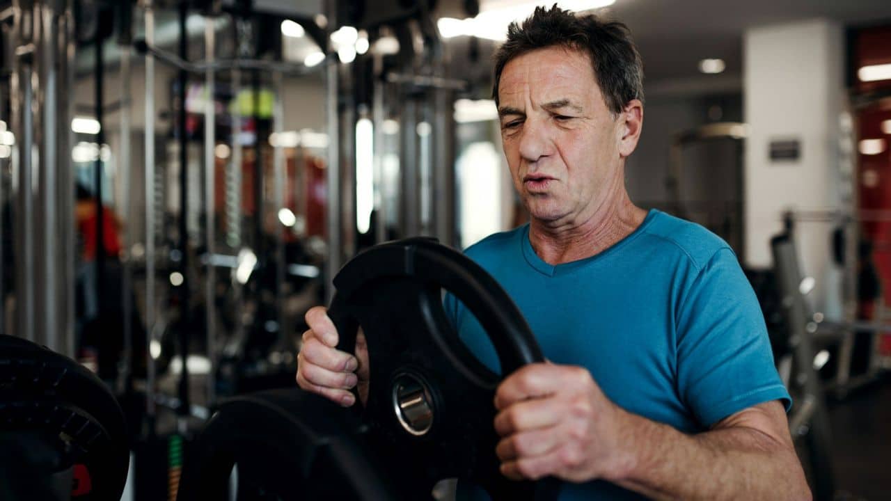 A focused older man in a blue shirt holds a black weight plate in a gym.