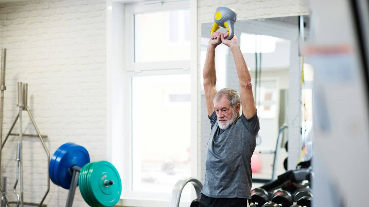 A determined senior man lifts a kettlebell over his head in a gym.
