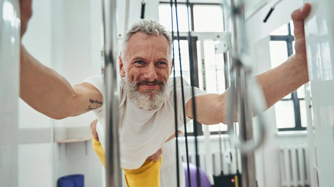 A joyful older man with a beard leans on a piece of gym equipment.