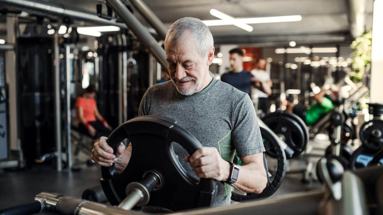 A serious older man with grey hair holds a weight plate while standing in a gym.