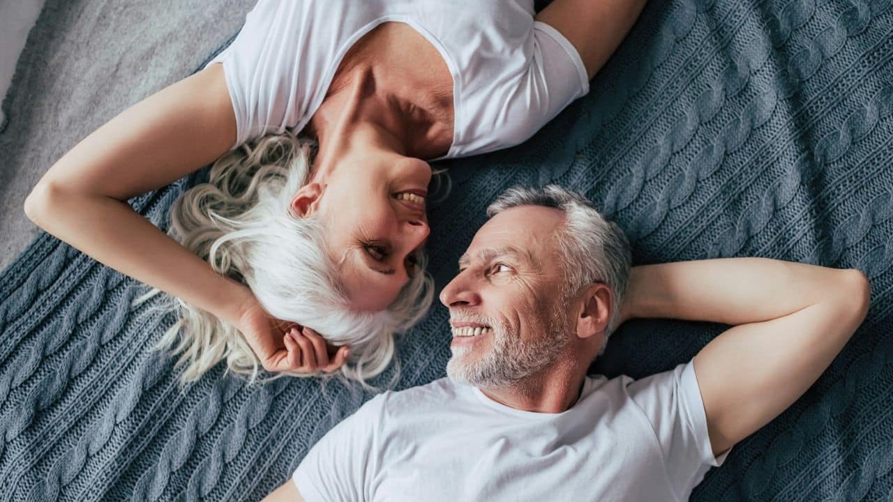 A happy, older couple with white hair lies in bed, looking at each other.