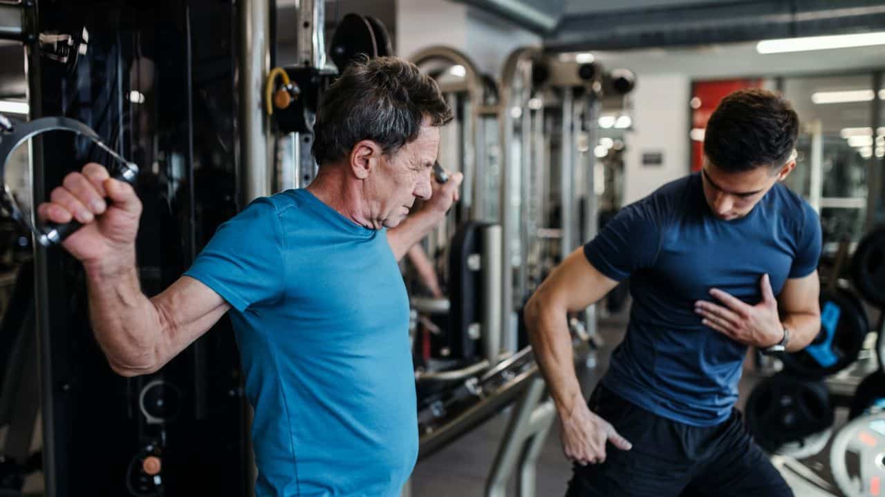 A serious older man uses a cable machine while a younger man assists him.