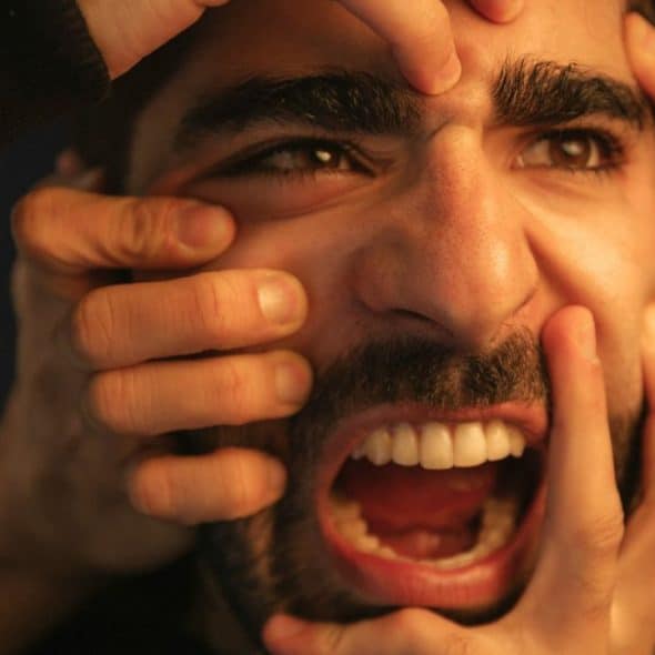 A stressed man with a beard and mustache screams as multiple hands touch his face.
