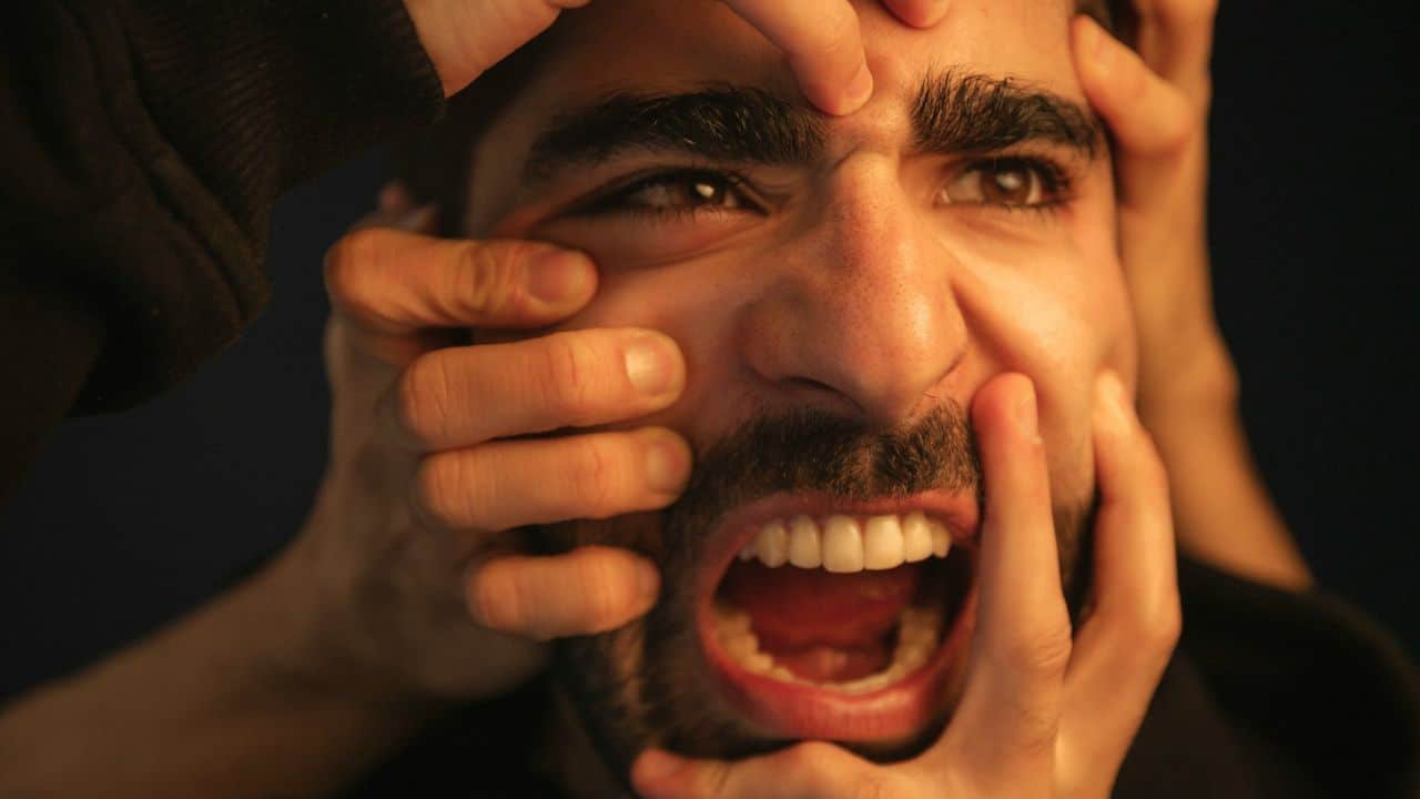 A stressed man with a beard and mustache screams as multiple hands touch his face.