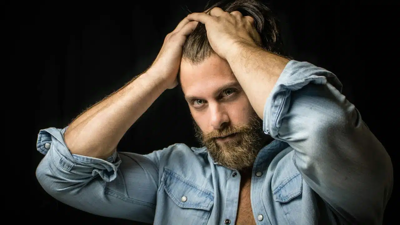 A serious, bearded man with a denim shirt holds his hair with both hands.