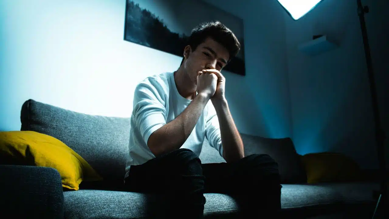 A young man sits on a couch with his hands clasped under his chin.