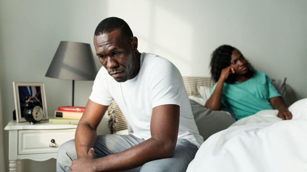 A man sits on the edge of a bed, looking upset, while a woman lies behind him.