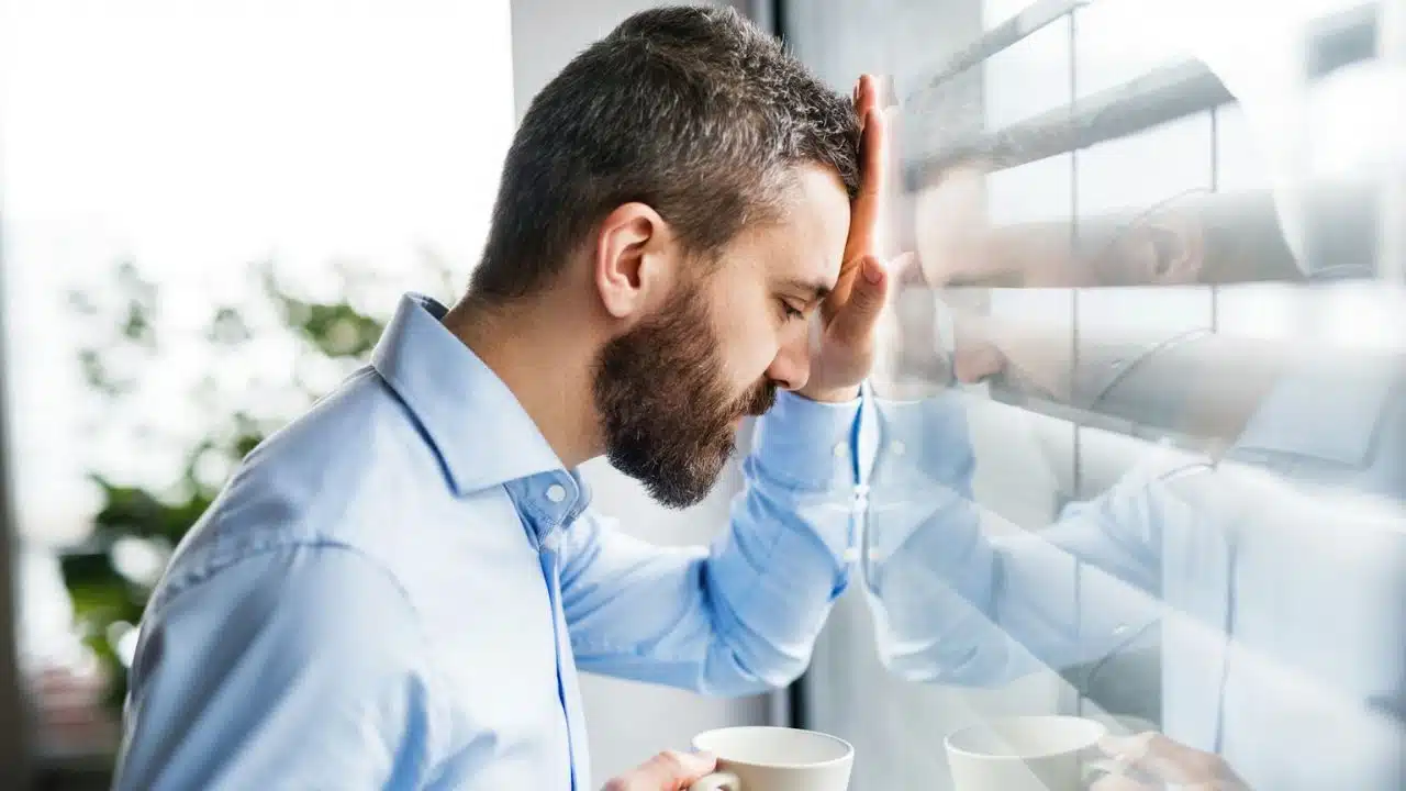 A bearded man leans his forehead against a window, holding a white mug.