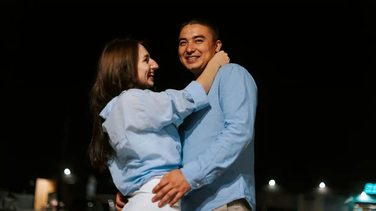 A smiling couple embraces outdoors at night, looking at each other.