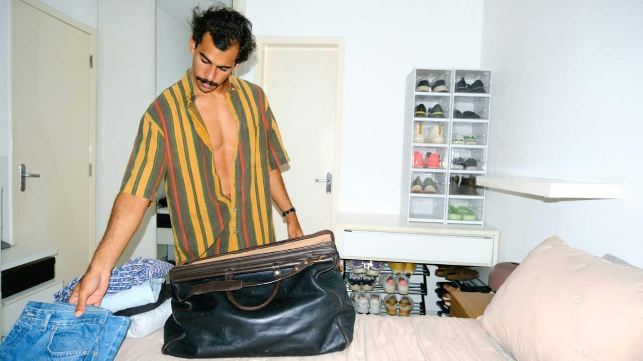 A man with a mustache packs clothes into a bag on a bed.