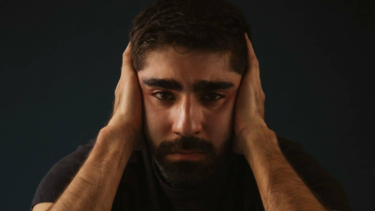 A distressed man covers his ears with his hands, looking at the camera.