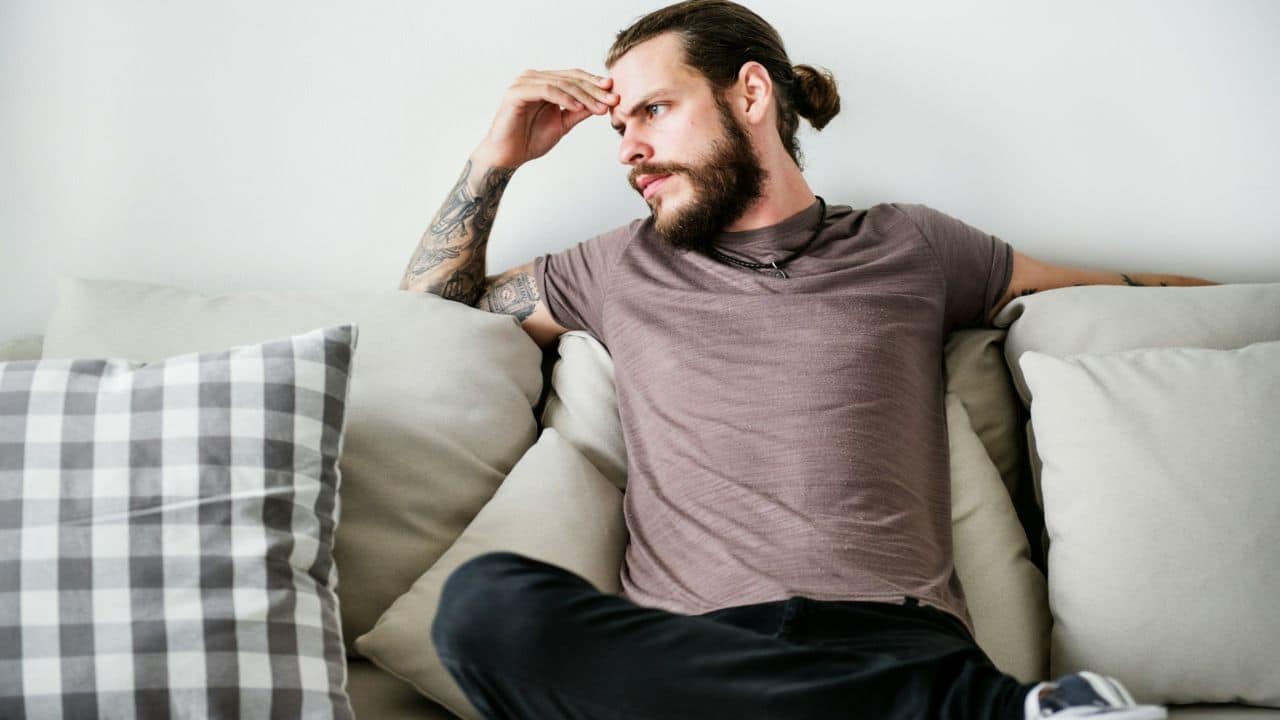 A man with a beard, man bun, and tattoos sits on a couch, looking away thoughtfully.