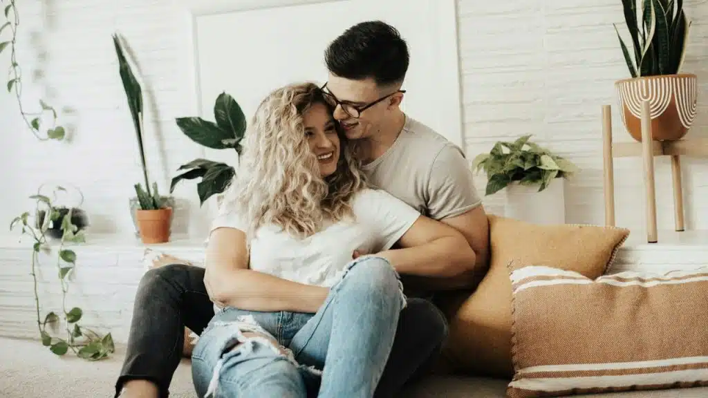 A happy couple embraces on the floor, surrounded by houseplants.