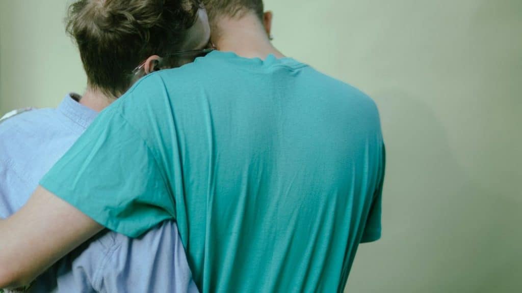 Two people embrace, seen from behind, with one person's head on the other's shoulder.