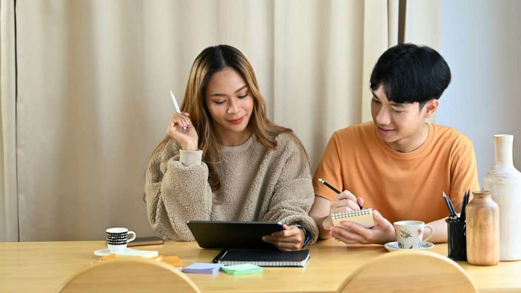A man and woman sit at a table, looking at a tablet and a notebook.