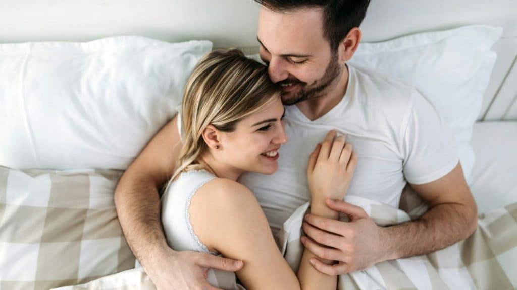 A happy couple lies in bed, embracing and smiling at each other.