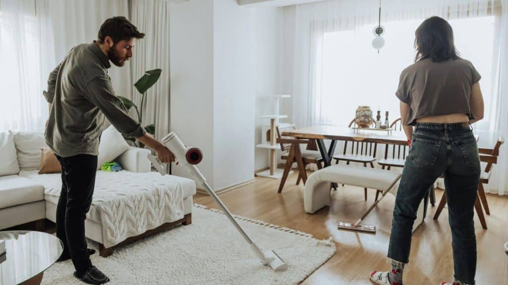 A man vacuums the living room while a woman stands in the background, facing away.