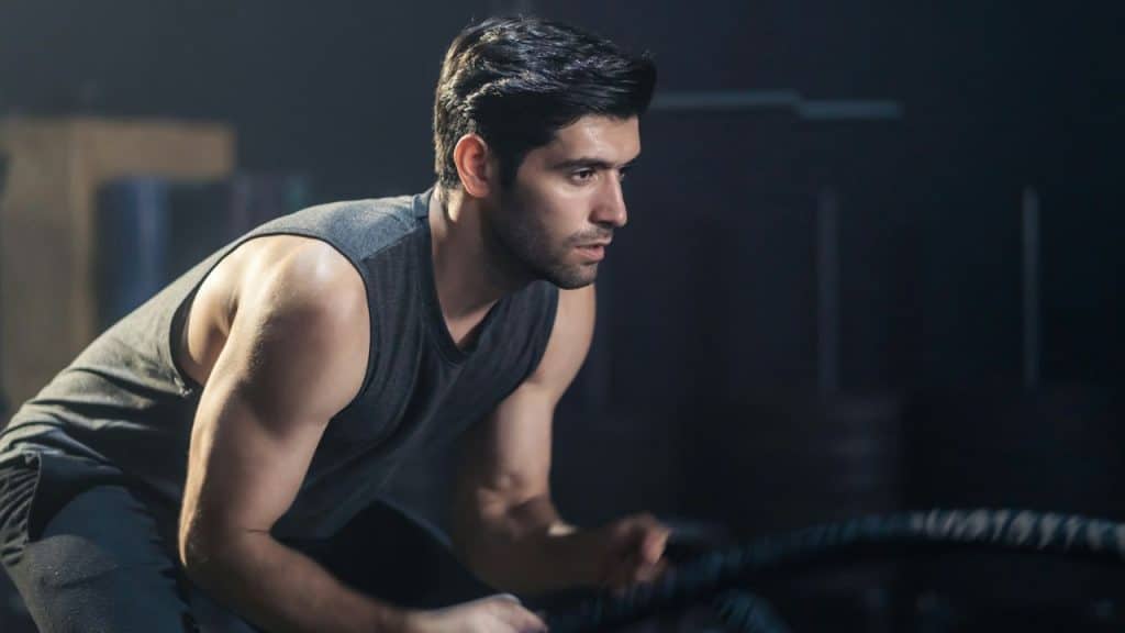 A focused man with dark hair works out with battle ropes at a gym.