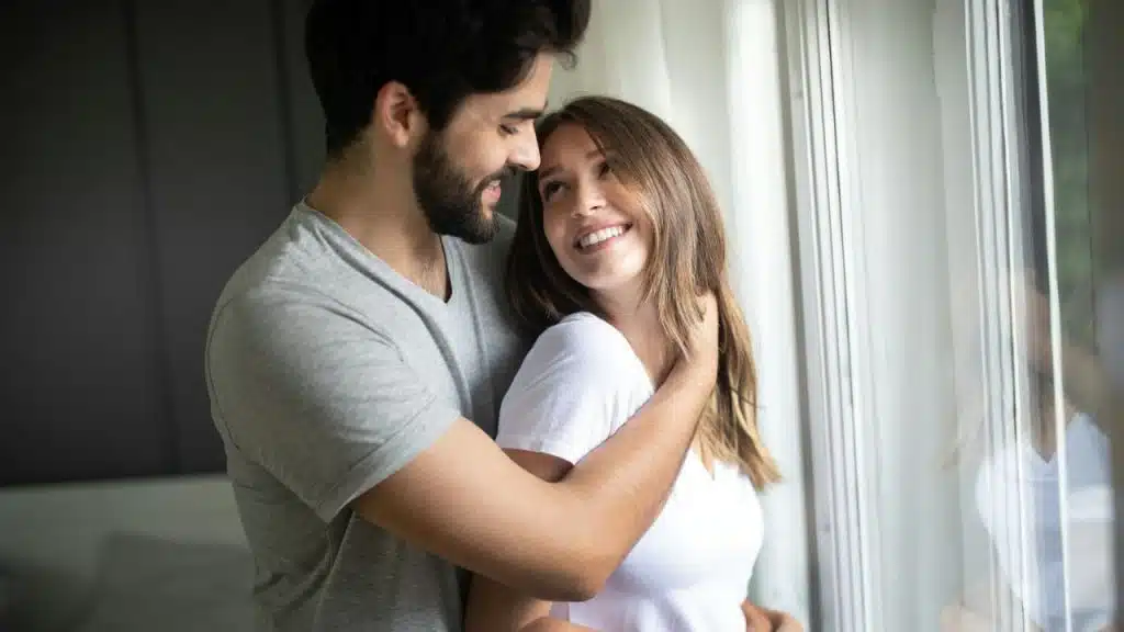 A smiling couple embraces while standing next to a window.