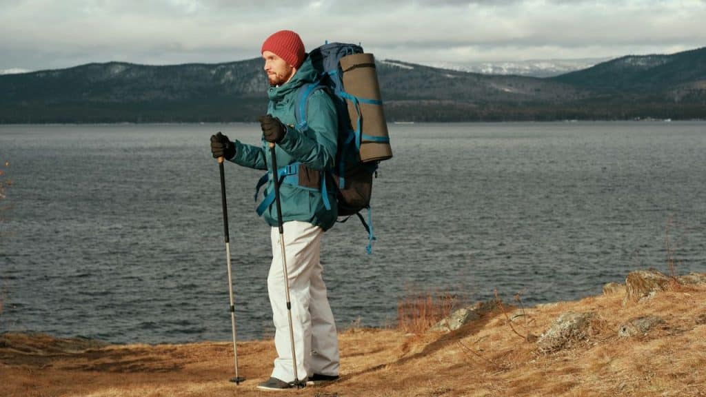A male hiker with a backpack and poles stands on a hill by a lake.