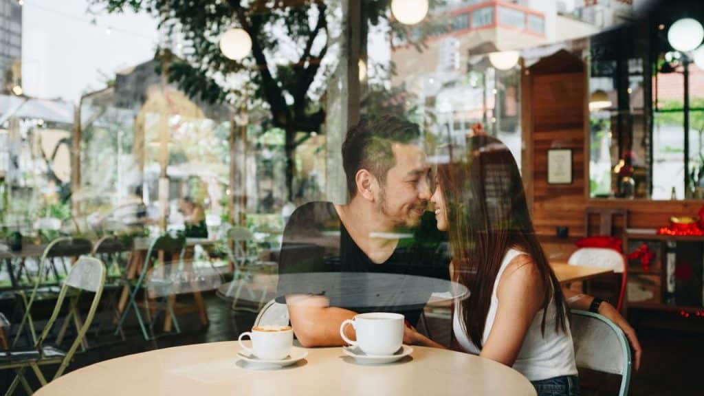 A happy couple smiles at each other at a cafe table with coffee.
