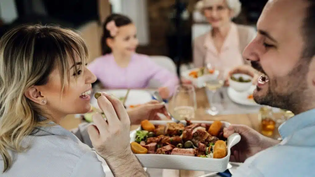 A couple smiles and shares food at a dinner table with family.