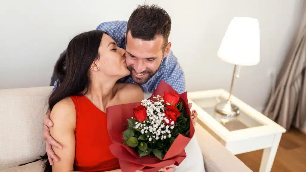 A smiling man gives a woman a bouquet of roses and kisses her on the cheek.