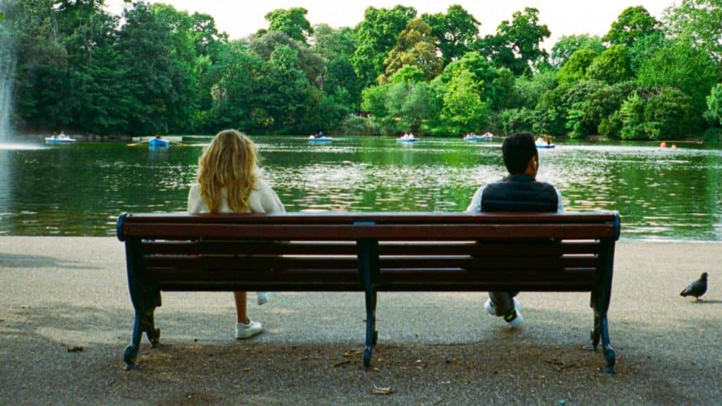 A woman and a man sit on a park bench facing a lake, not looking at each other.