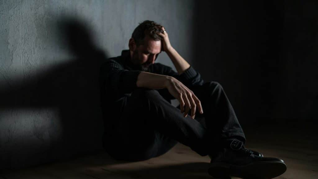 A stressed man sits against a wall, holding his head with his hand and looking down.