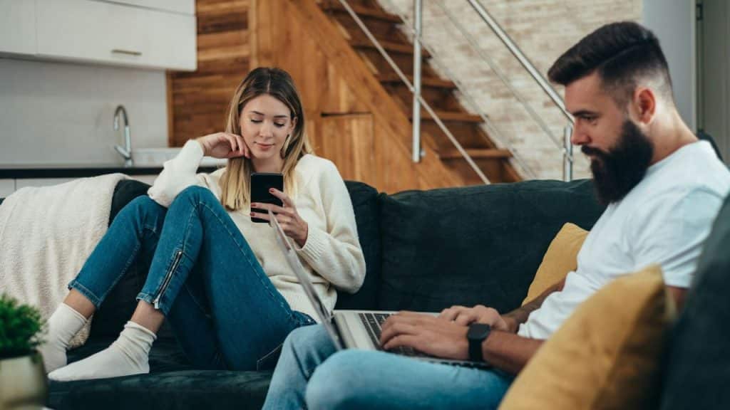 A woman looks at her phone while a man next to her uses a laptop on a couch.