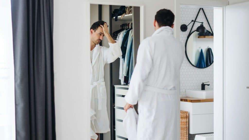 A man in a white robe looks in a mirror, adjusting his hair in a closet.