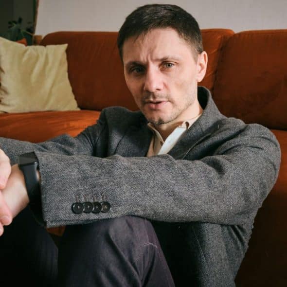 A man in a gray jacket and a watch sits on the floor, looking at the camera.